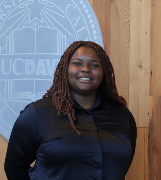 Headshot photo of smiling woman with braids, dark shirt, UC Davis seal on wood wall