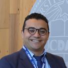 Smiling man in glasses and suit with blue lanyard, wooden wall and circular logo behind