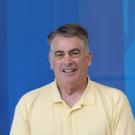 Smiling middle-aged man in yellow polo, portrait against blue geometric background.