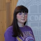 Young woman with glasses, purple sweatshirt, wood wall and round emblem behind