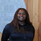 Headshot photo of smiling woman with braids, dark shirt, UC Davis seal on wood wall